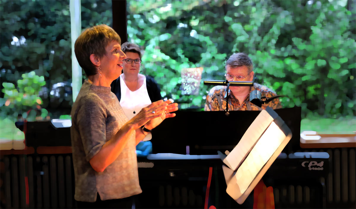 Claudia Michel im Profil singend im Vordergrund links, mit warmen Licht angestrahlt von der Notenst�nderbeleuchtung rechts. Im Mittelgrund dahinter enface Annegret L�tteken beobachtend und Michael Herberts klavierspielend hinter dem Stagepiano. Der Hintergrund zeigt durch die gro�e Scheibenfront des Thomaszentrums die saftgr�n belaubten B�ume und Str�ucher.